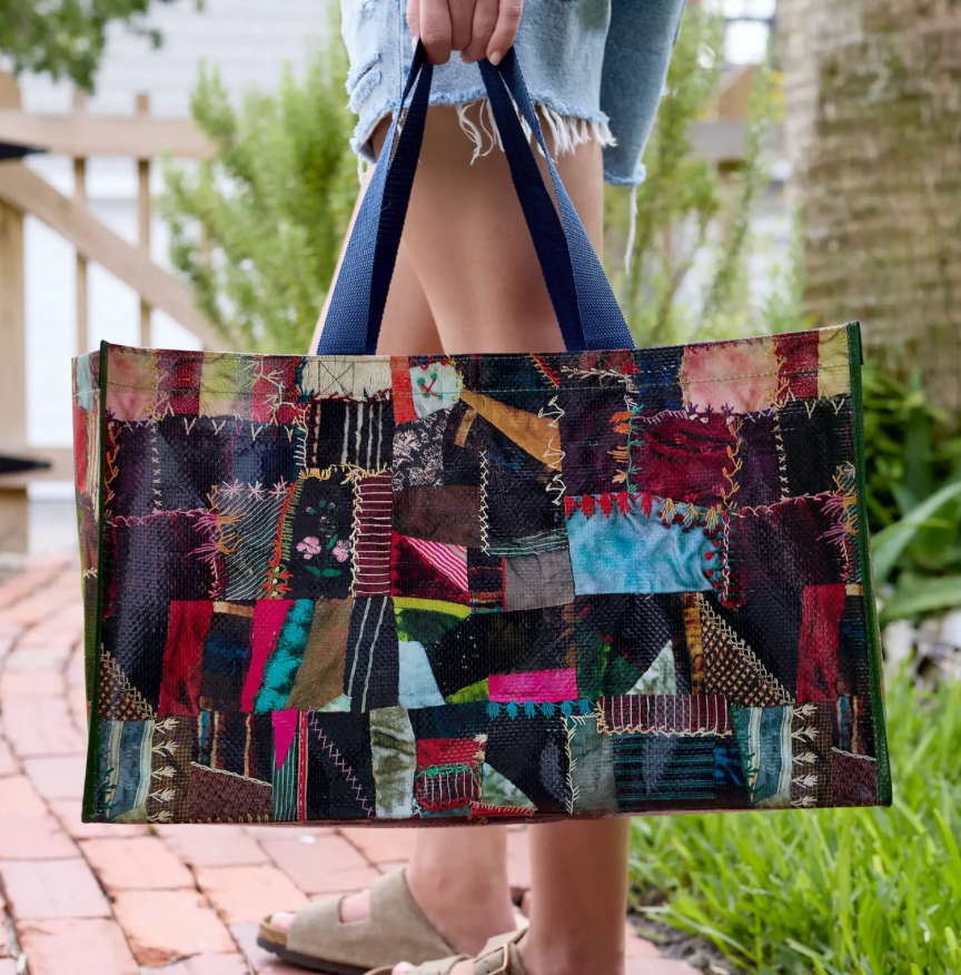 A person outdoors holding a large patchwork fabric tote bag with dark blue handles. The bag features a colorful quilted patchwork pattern with various textile panels in red, green, blue, black, and pink.
