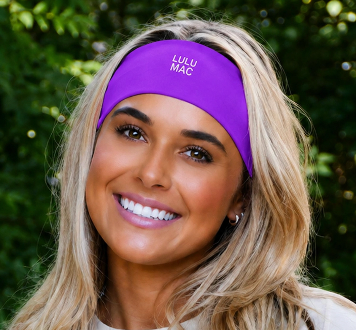 A smiling blonde woman wearing a bright purple athletic headband with the text 'LULU MAC' printed in white on the front, set against a blurred green outdoor background.