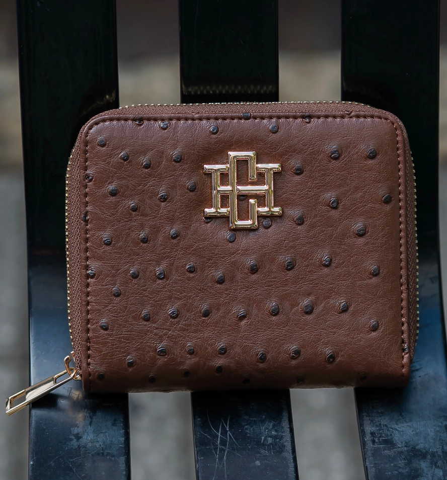 A brown ostrich-textured leather zip-around wallet with a gold-tone interlocking H logo on the front, resting on a black slatted bench.