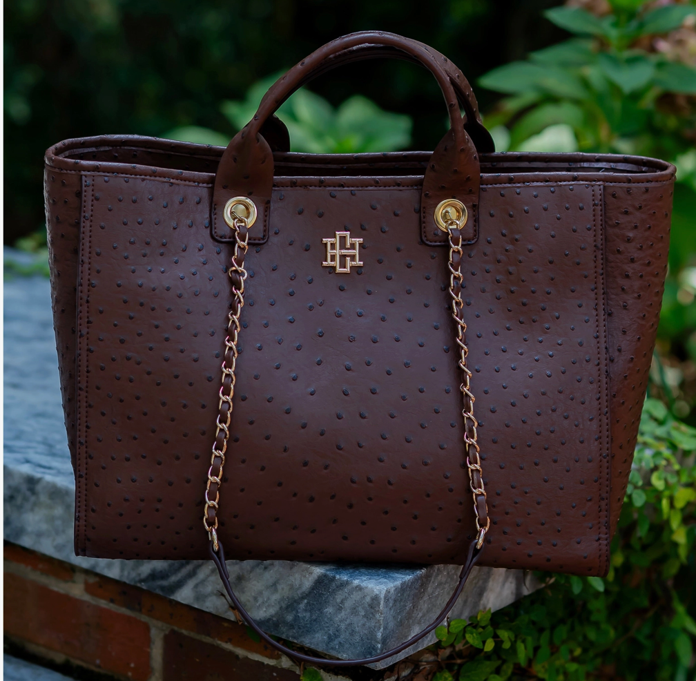 A dark brown ostrich-textured leather tote bag with gold-tone hardware and a central logo, resting on a stone ledge with green foliage in the background.