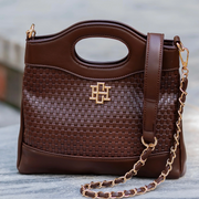 A brown woven leather handbag with a top handle, a gold-tone interlocking monogram logo on the front, and a matching brown leather shoulder strap attached to a gold chain, shown against a blurred outdoor background.