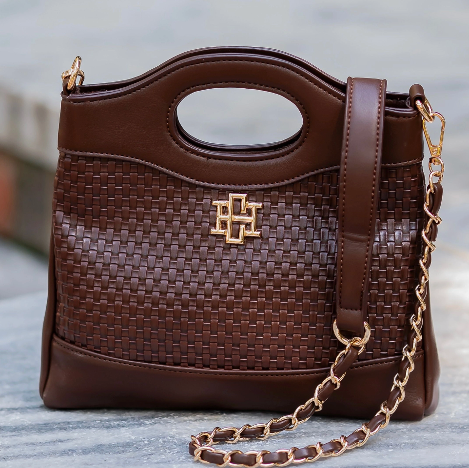 A brown woven leather handbag with a top handle, a gold-tone interlocking monogram logo on the front, and a matching brown leather shoulder strap attached to a gold chain, shown against a blurred outdoor background.