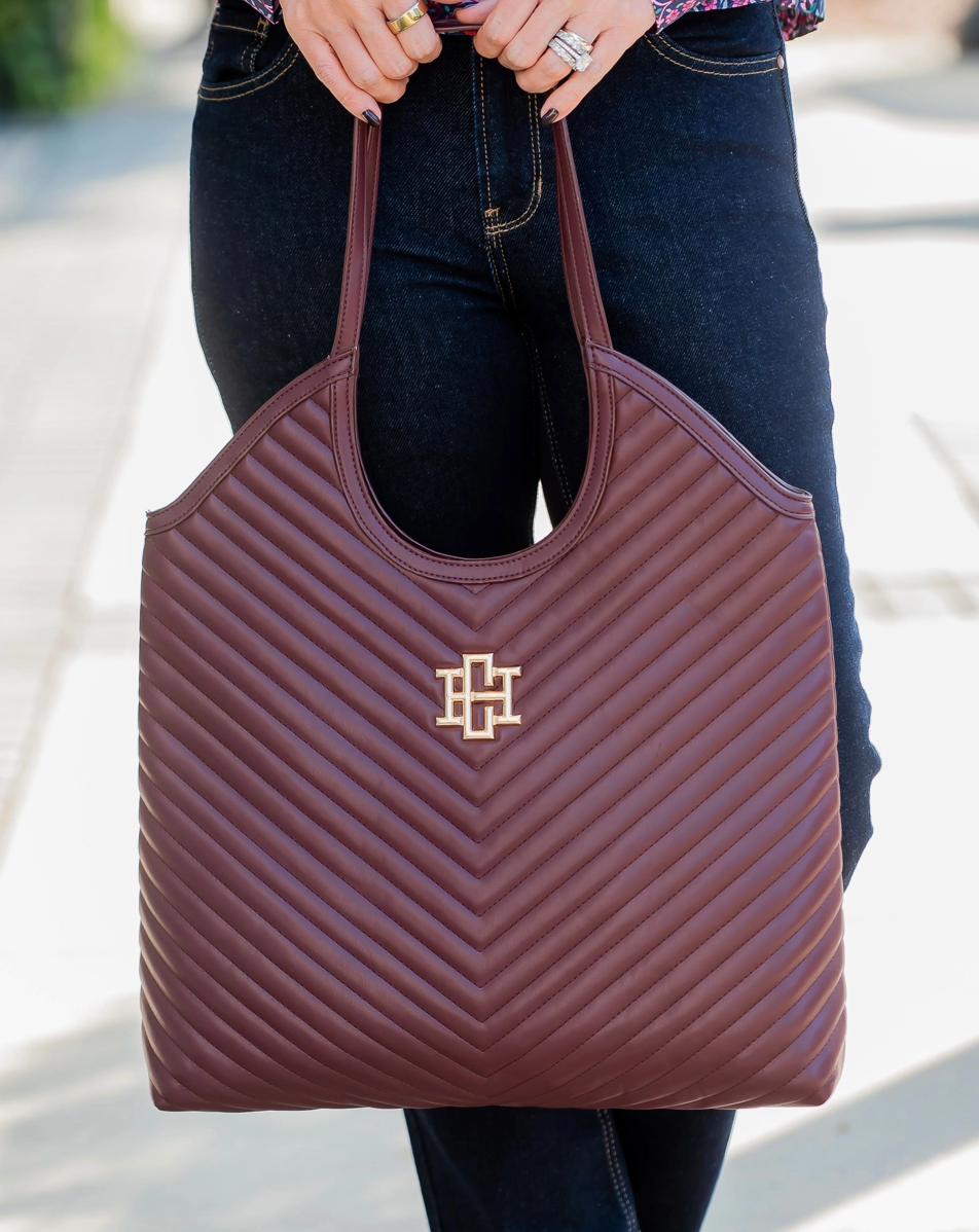 A person wearing dark blue jeans holds a large quilted burgundy tote bag with a gold interlocking monogram logo on the front. The photo is taken outdoors on a sunlit paved walkway.