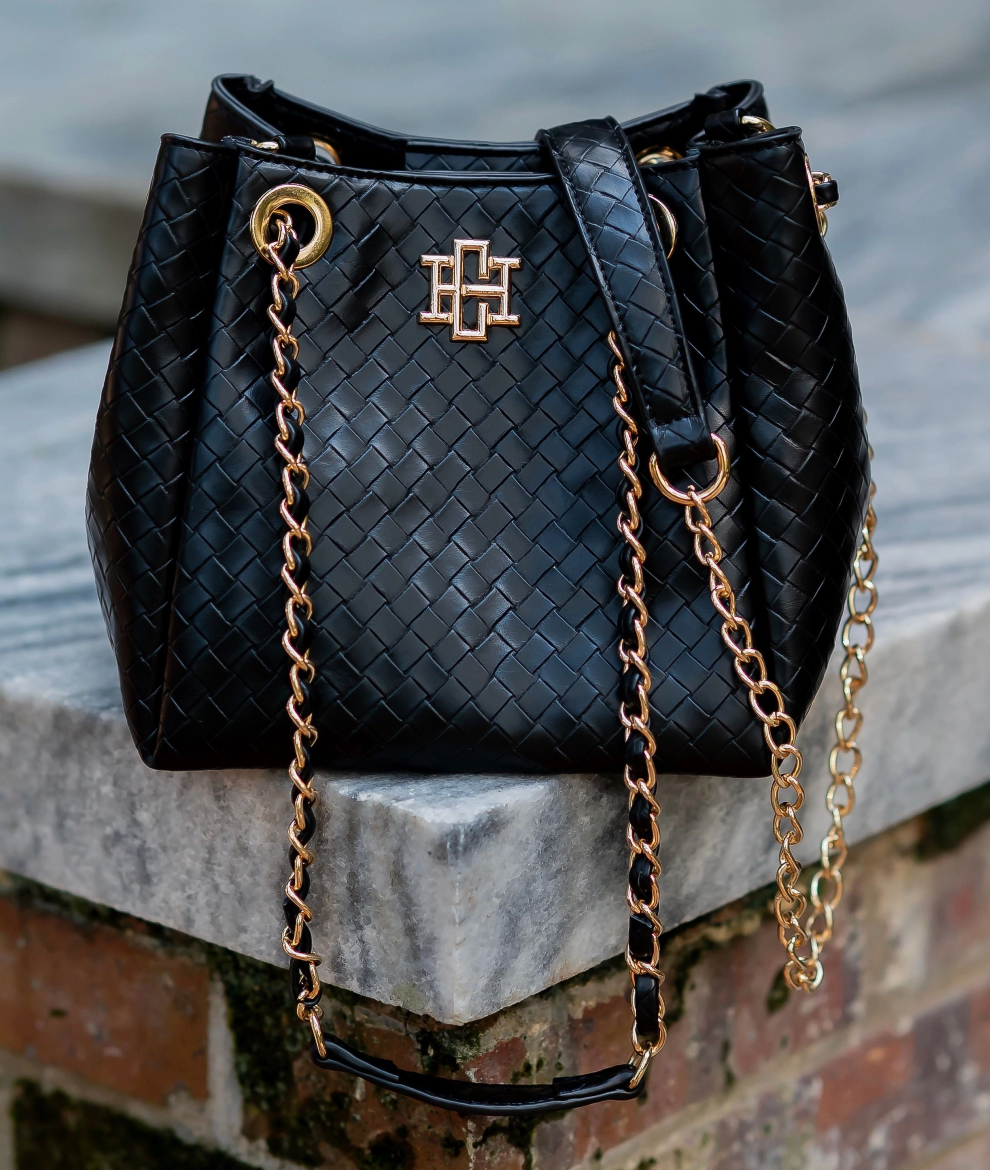 A black woven leather bucket bag with gold-tone hardware and a cross logo on the front, resting on a stone ledge outdoors.