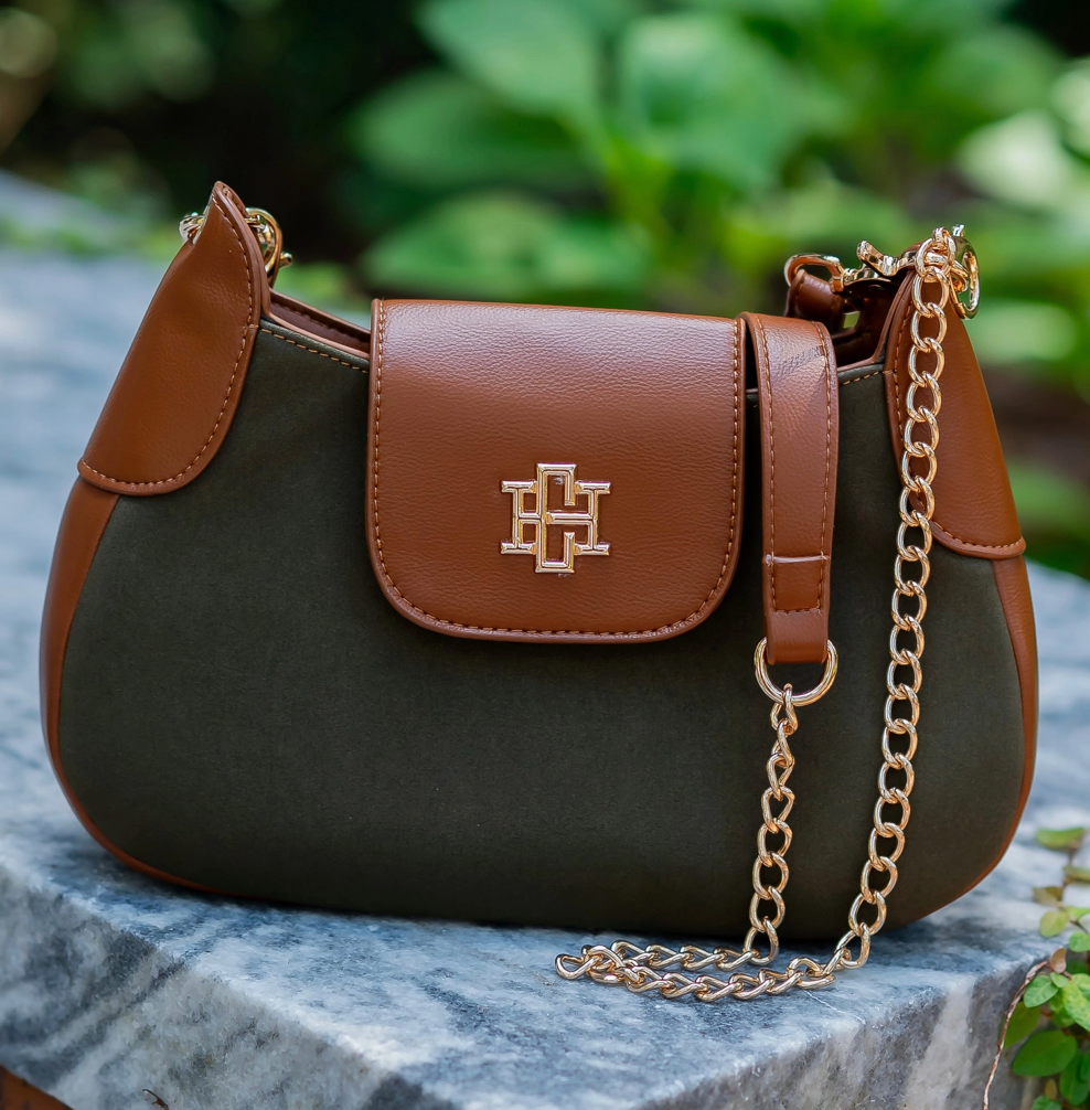 A structured shoulder bag with a dark olive green body and tan leather trim, featuring a gold-tone interlocking monogram logo on the front flap and a gold chain strap, resting on a gray stone surface with a blurred green garden background.