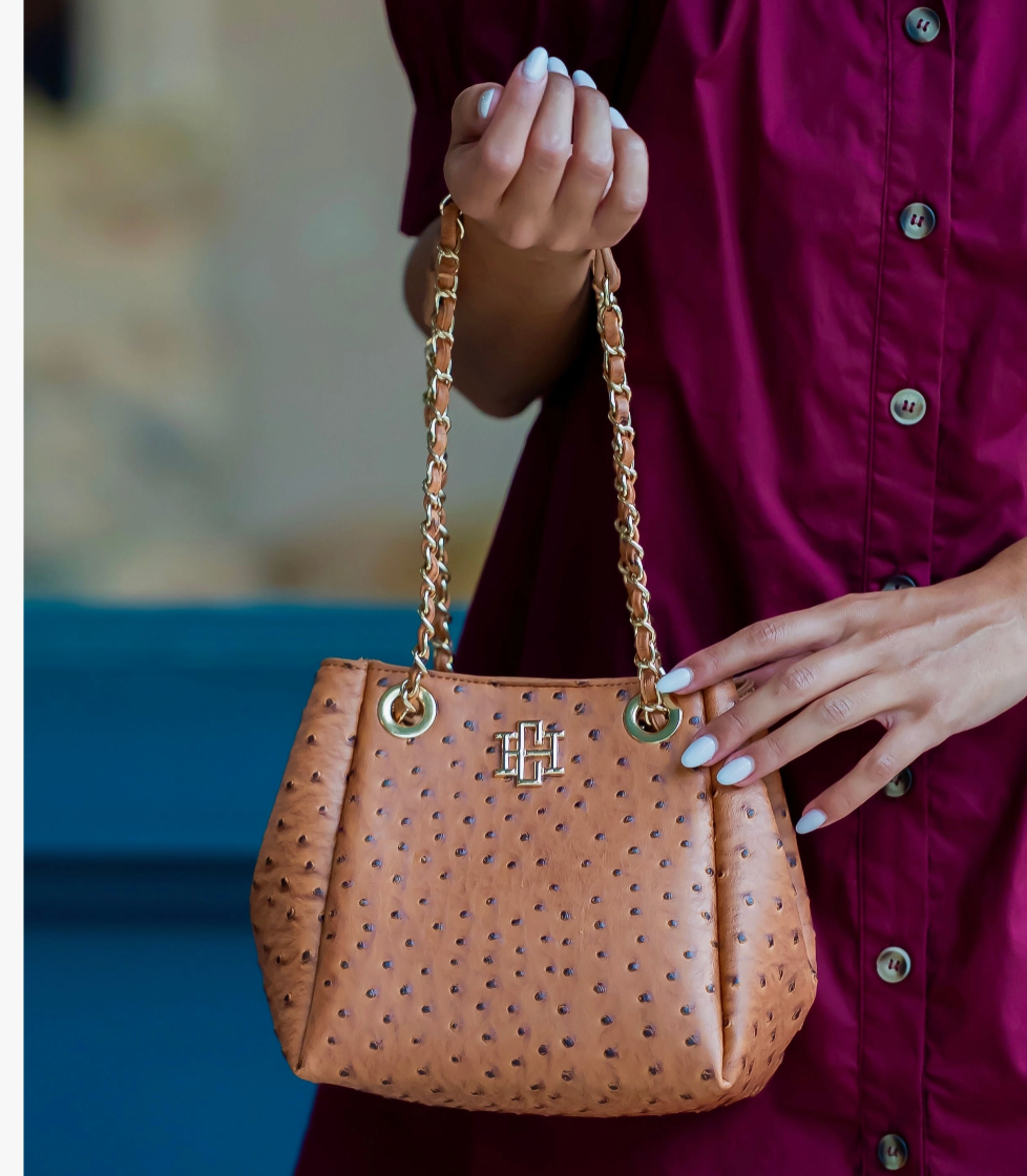 A tan ostrich-embossed leather handbag with gold-tone chain handles and a central monogram logo, held by a person wearing a burgundy button-up shirt.