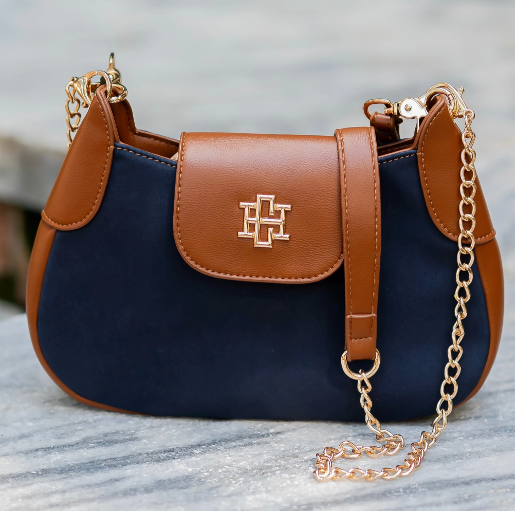 A navy blue and brown leather shoulder bag with a gold-tone cross logo on the front flap and a gold chain strap, resting on a gray marble surface.