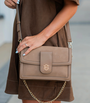 A taupe brown suede crossbody handbag with a gold-tone logo clasp and chain strap, carried by a model wearing a matching brown dress.