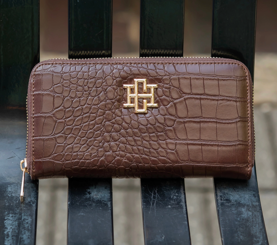 A brown crocodile-embossed zip-around wallet with a gold-tone interlocking H logo on the front, resting on a dark slatted bench.