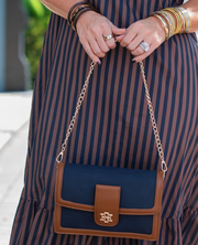 A navy blue and brown crossbody handbag with a gold chain strap, held by a person wearing a navy and brown vertically striped dress and gold bangles.