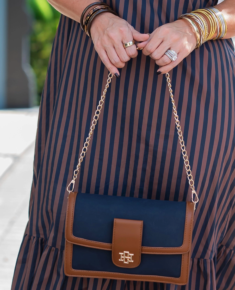 A navy blue and brown crossbody handbag with a gold chain strap, held by a person wearing a navy and brown vertically striped dress and gold bangles.