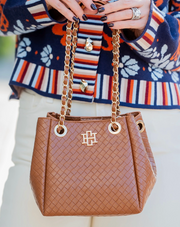 A brown woven leather handbag with gold-tone chain handles and a gold monogram logo, held by a person wearing a colorful striped cardigan and white pants.