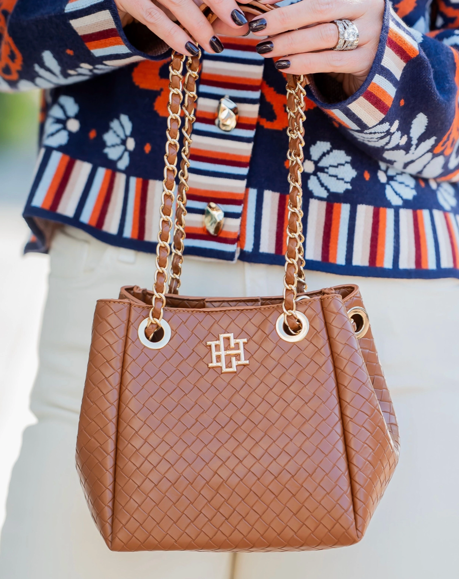 A brown woven leather handbag with gold-tone chain handles and a gold monogram logo, held by a person wearing a colorful striped cardigan and white pants.
