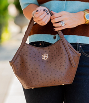 A brown ostrich-textured handbag with a gold monogram logo, held by a person wearing a turquoise and brown cardigan, black jeans, and rings.