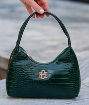 A dark green crocodile-embossed leather handbag with a gold-tone interlocking logo clasp, held by a hand with white manicured nails against a blurred gray stone background.
