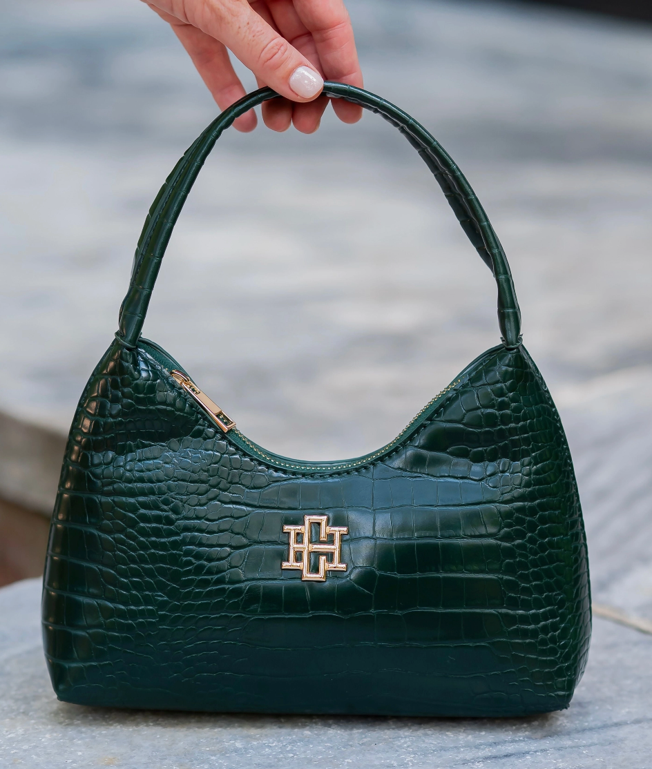 A dark green crocodile-embossed leather handbag with a gold-tone interlocking logo clasp, held by a hand with white manicured nails against a blurred gray stone background.