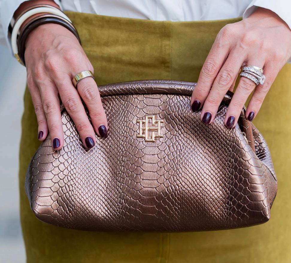 A close-up of a person holding a metallic bronze textured handbag with a gold interlocking logo on the front. The person is wearing a mustard yellow top and multiple bracelets and rings.