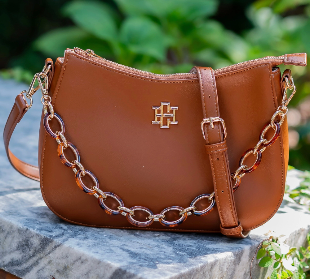 A brown leather shoulder bag with a gold-tone monogram logo and a chunky chain strap, resting on a gray stone surface with green foliage in the background.