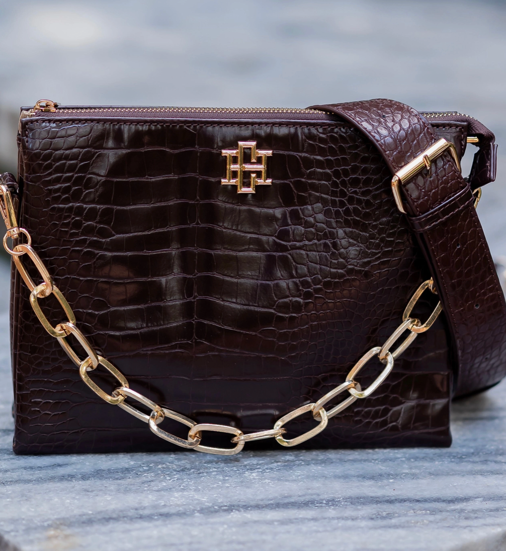 A dark brown crocodile-embossed leather handbag with a gold-tone chain strap and a gold monogram logo on the front, displayed on a gray stone surface.