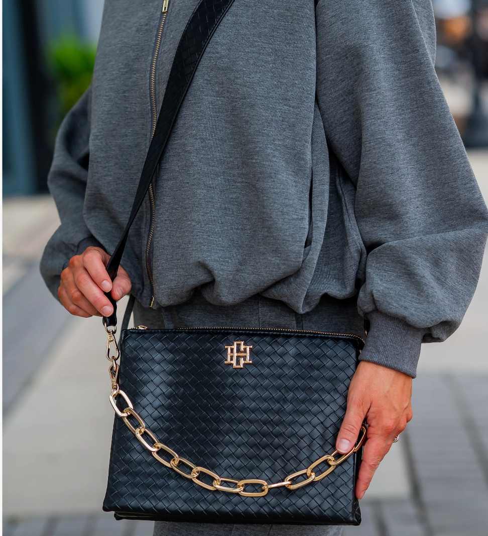 A person wearing a gray sweatshirt carries a black woven crossbody bag with a gold chain detail and a gold monogram logo on the front, photographed outdoors on a blurred street background.
