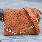 A brown ostrich-embossed leather crossbody bag with a gold-tone chain strap and a gold cross logo on the front, resting on a gray stone surface.