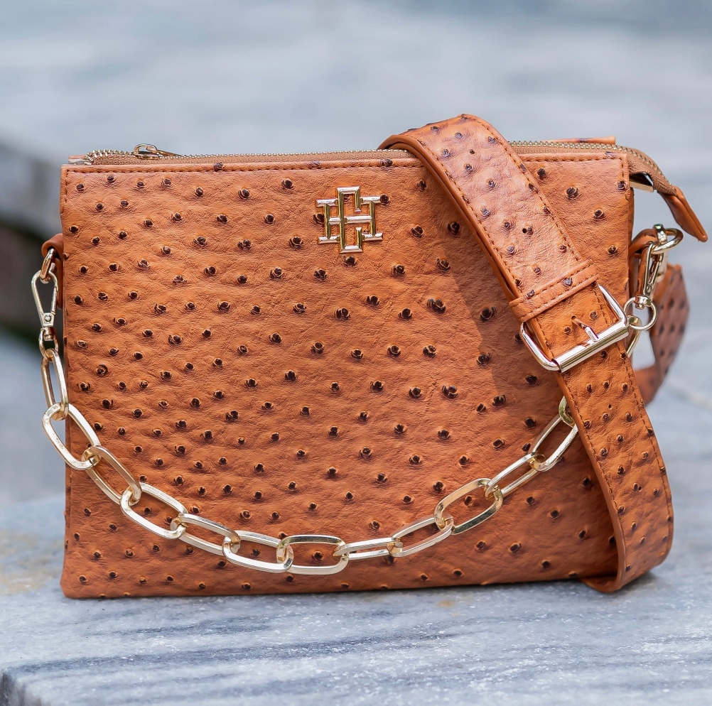A brown ostrich-embossed leather crossbody bag with a gold-tone chain strap and a gold cross logo on the front, resting on a gray stone surface.