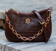 A dark brown crocodile-embossed leather handbag with a gold-tone chain strap and a central gold monogram logo, resting on a gray stone surface.