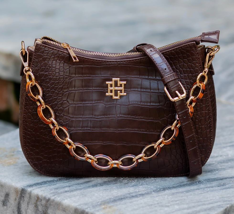 A dark brown crocodile-embossed leather handbag with a gold-tone chain strap and a central gold monogram logo, resting on a gray stone surface.