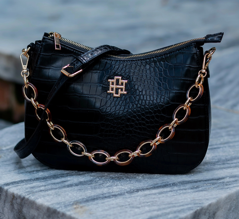 Black crocodile-embossed leather handbag with a gold-tone chain strap and a central cross logo, resting on a gray stone surface.