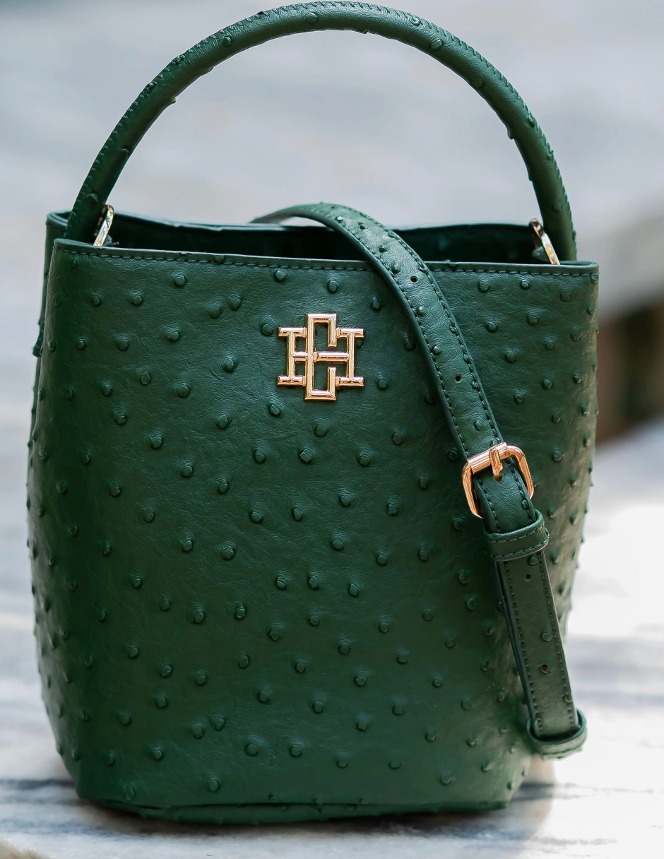 A dark green ostrich leather bucket bag with a top handle, adjustable shoulder strap, and a gold-tone interlocking monogram logo on the front, set against a blurred outdoor background.