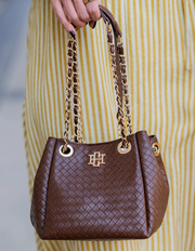 A brown woven leather handbag with gold-tone chain straps and a gold monogram logo, held by a person wearing a yellow and white striped dress.