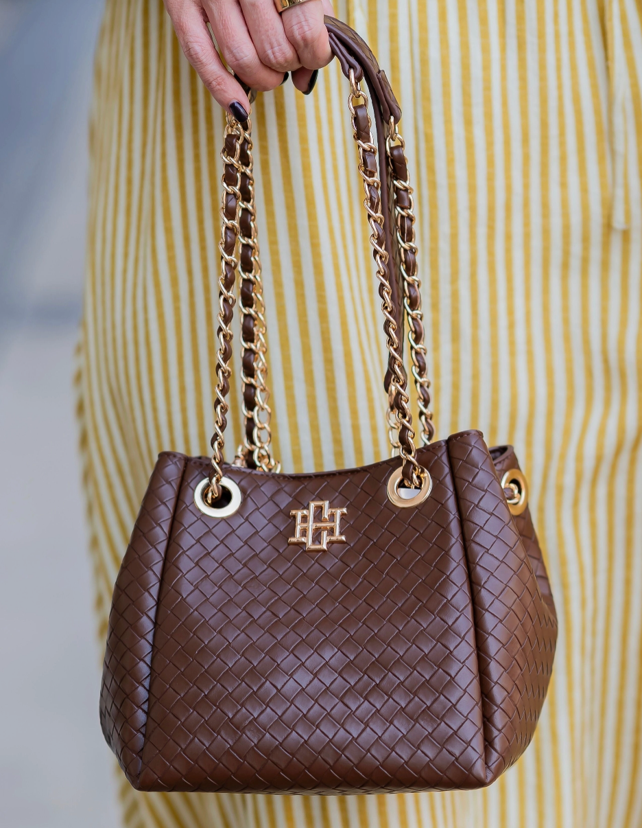A brown woven leather handbag with gold-tone chain straps and a gold monogram logo, held by a person wearing a yellow and white striped dress.