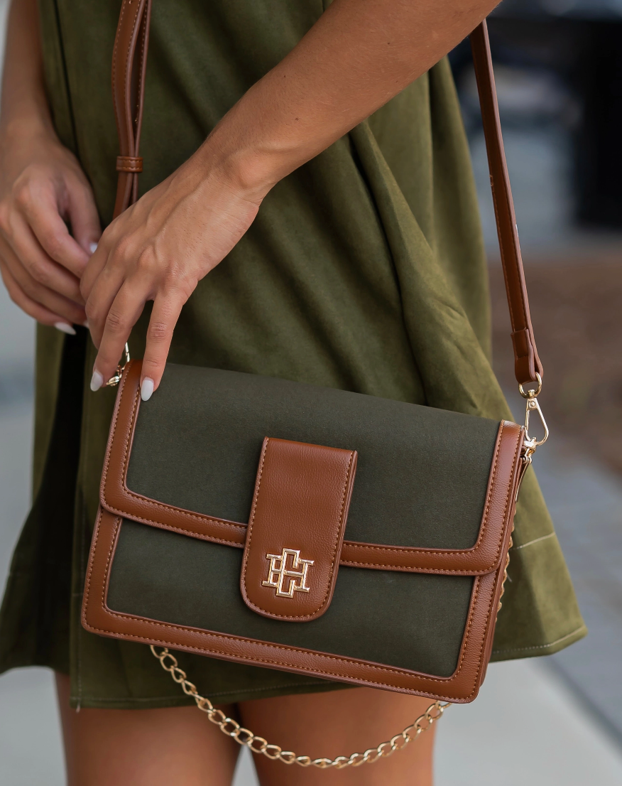 An olive green crossbody bag with brown leather trim and a gold-tone logo clasp, carried by a model wearing an olive green dress.