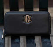 A black leather zip-around wallet with a prominent gold-tone interlocking 'H' logo on the front, resting on a dark wooden slatted bench.
