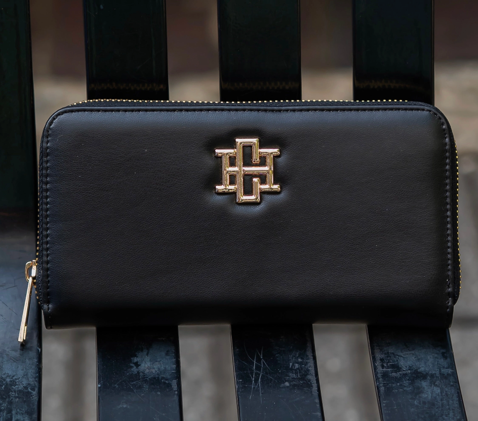 A black leather zip-around wallet with a prominent gold-tone interlocking 'H' logo on the front, resting on a dark wooden slatted bench.