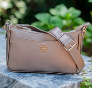 A taupe pebbled leather shoulder bag with a gold-tone interlocking monogram logo, resting on a gray marble surface with a blurred green garden background.