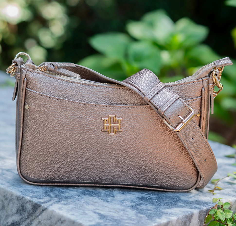 A taupe pebbled leather shoulder bag with a gold-tone interlocking monogram logo, resting on a gray marble surface with a blurred green garden background.