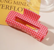 A red and white gingham check hair claw clip with white lettering that reads 'I LOVE CHINA' along the side, resting on a yellow book with a glass of water in the background.