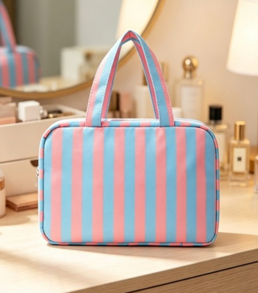 A rectangular zippered cosmetic bag with vertical pink and light blue stripes, featuring two top handles, displayed on a wooden vanity table in a softly lit room.