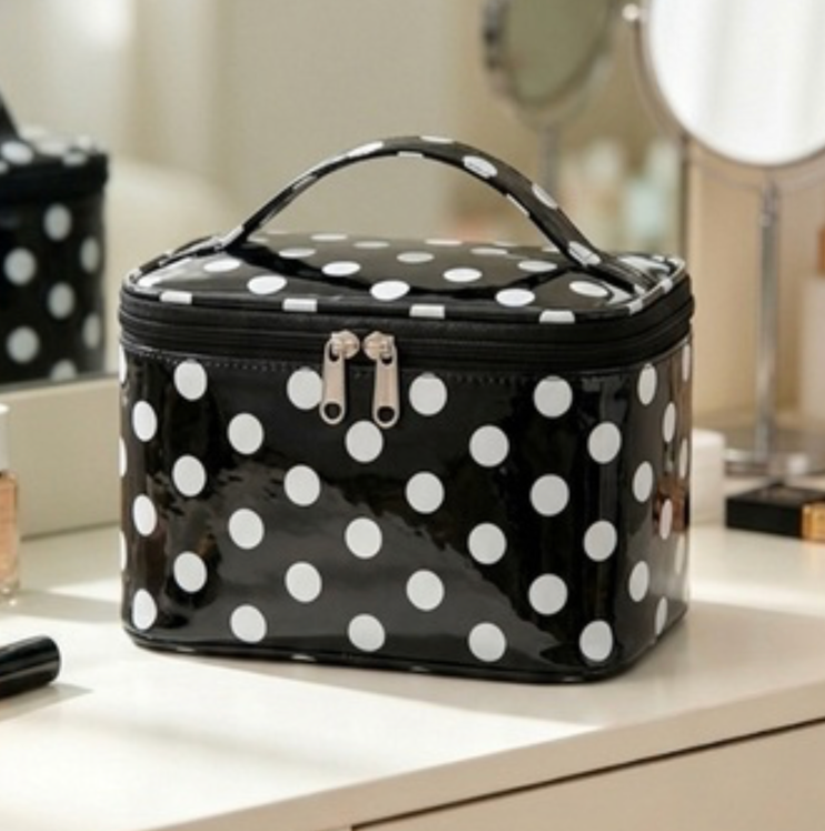 A glossy black cosmetic bag with white polka dots sits on a white vanity table in a softly lit room. The bag has a top handle, a zip closure with silver-toned hardware, and is shown alongside other vanity items in the background.