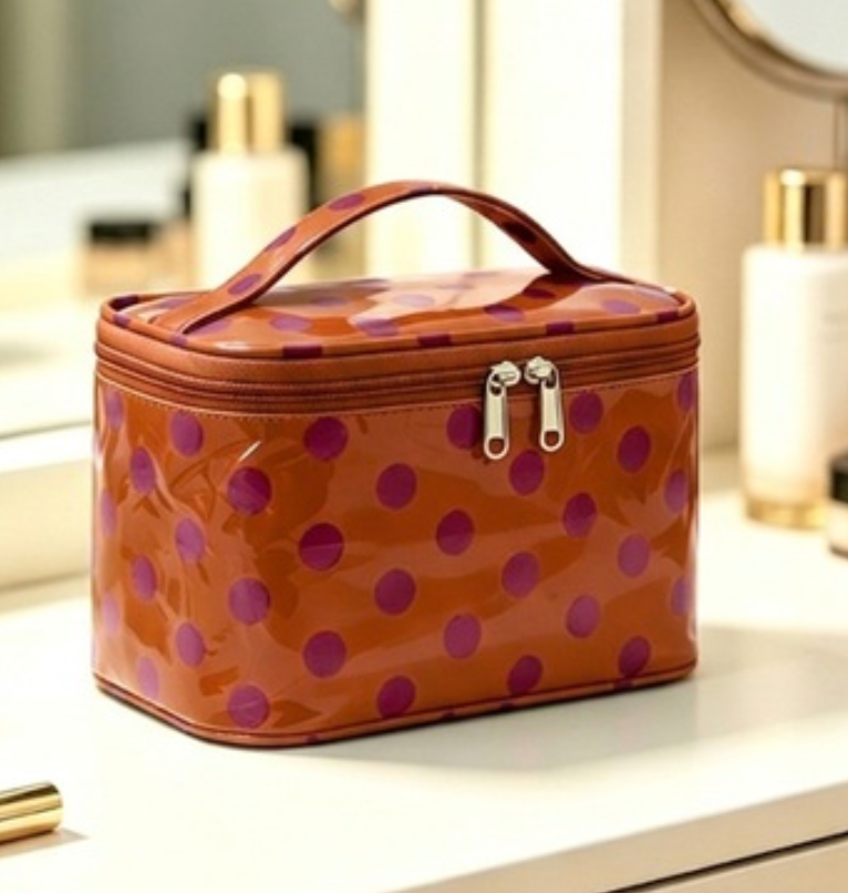 A glossy orange cosmetic bag with purple polka dots, featuring a top handle and dual silver zipper pulls, resting on a white vanity counter with blurred skincare bottles in the background.