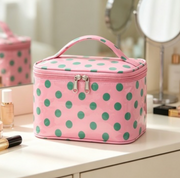 Pink cosmetic bag with green polka dots, featuring a top handle and dual zipper pulls, displayed on a white vanity table with makeup items and mirrors in the background.
