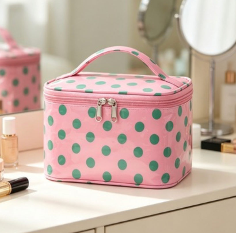 Pink cosmetic bag with green polka dots, featuring a top handle and dual zipper pulls, displayed on a white vanity table with makeup items and mirrors in the background.