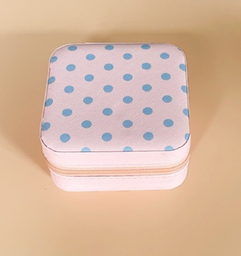 A square zippered jewelry box with a light pink textured fabric cover decorated with blue polka dots, set against a solid beige background.