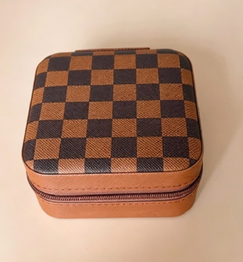A square zippered case with a brown and black checkerboard pattern and tan leather trim, photographed on a plain light beige background.