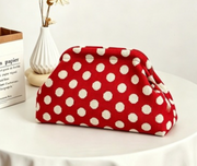 A red fabric pouch with white polka dots sits on a white table next to a small white vase with dried stems and a book.