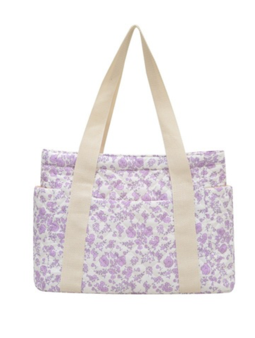 A fabric tote bag with a light purple floral print on a white background, featuring two thick beige shoulder straps and a front pocket.