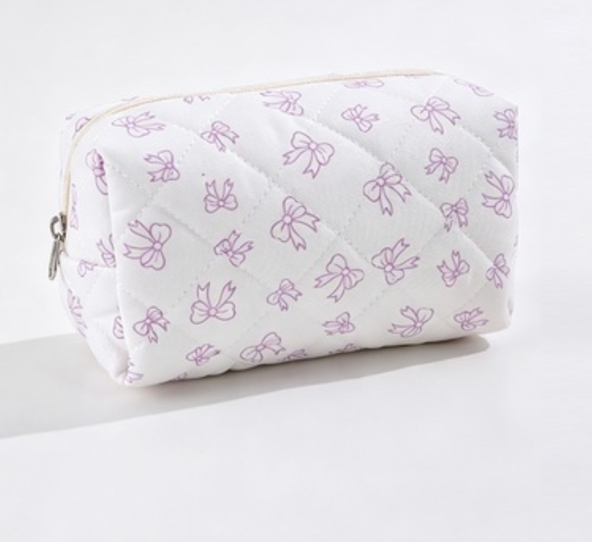 A quilted cosmetic pouch with a white background and a repeating pattern of light purple bow outlines, featuring a silver zipper pull on the left side.