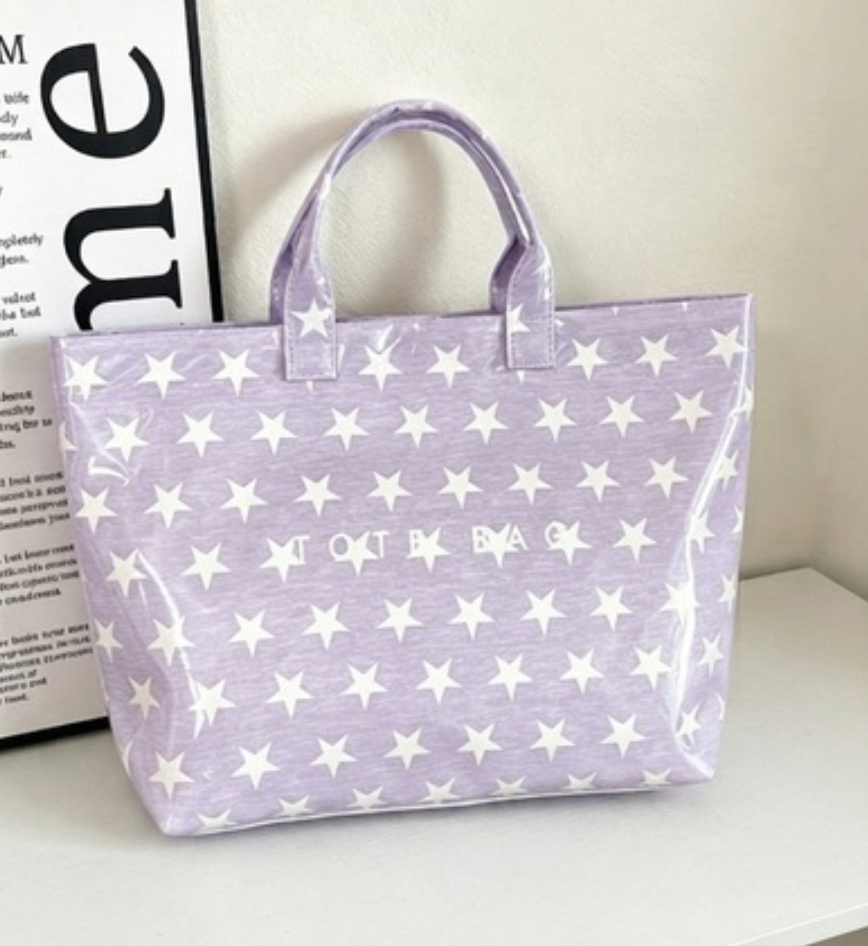 A lavender tote bag with a white star pattern and 'LOVE BAG' printed on the front, resting on a white surface next to a decorative book.