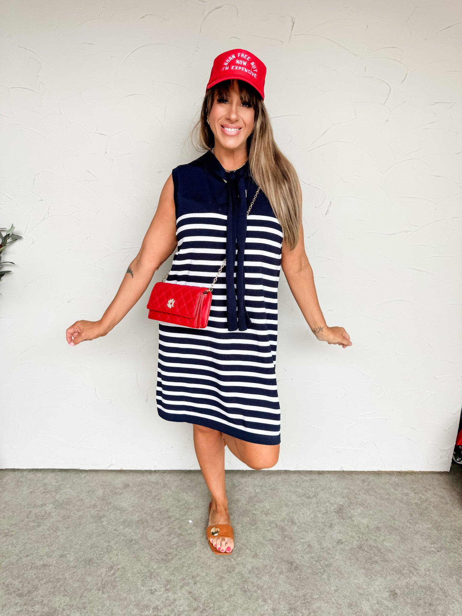 A woman smiling and standing on one leg against a textured off-white wall, wearing a sleeveless shift knit dress crafted from a 63% viscose and 37% nylon blend. The dress features a solid navy blue top section with a pointed collar and long, matching navy knit neck-tie straps that hang loosely down the front, transitioning into a navy and white horizontally striped pattern through the body. She is styled with a vibrant red trucker hat that reads "BORN FREE BUT NOW I'M EXPENSIVE", a quilted red crossbody han